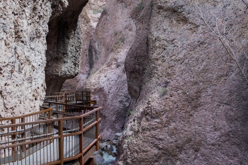 A winding wooden walkway through a narrow canyon with steep, rocky walls and a small stream below.