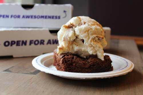 A brownie topped with a scoop of ice cream on a paper plate, with pizza boxes in the background.