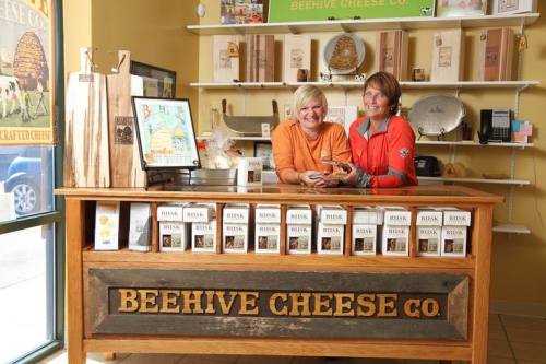 Two women smile behind the counter of Beehive Cheese Co., surrounded by cheese products and wooden displays.