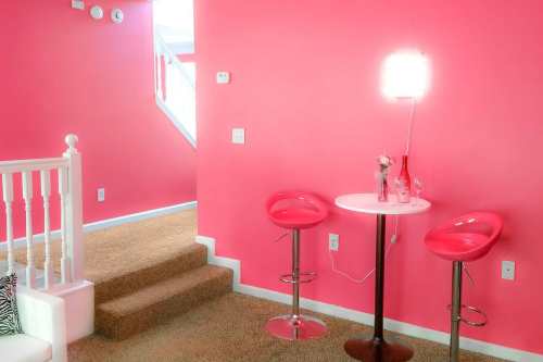 Bright pink walls with a small round table and two pink bar stools, leading to a staircase in a cozy living space.