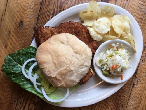 A plate with a breaded chicken sandwich, lettuce, onion, potato chips, and a side of coleslaw.