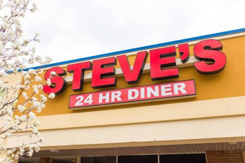 Sign for Steve's 24 Hour Diner, featuring bold red letters against a yellow background, with blooming flowers nearby.