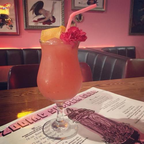A colorful cocktail with a lemon slice and flower garnish, served in a tall glass on a wooden table with a menu.