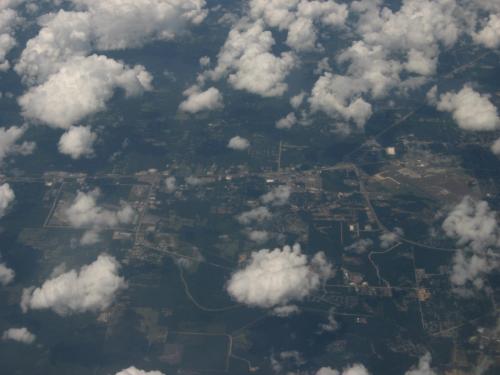 Aerial view of a landscape with patches of green fields, roads, and scattered clouds above.