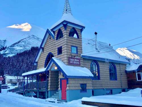 A charming, snow-covered art gallery with a tall spire, surrounded by mountains and trees under a clear blue sky.
