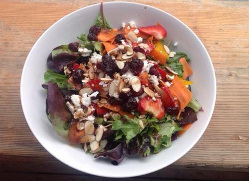 A colorful salad with mixed greens, strawberries, peppers, nuts, and feta cheese in a white bowl on a wooden surface.