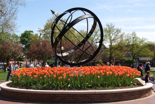 A large sundial surrounded by vibrant orange tulips in a sunny park with people enjoying the day.