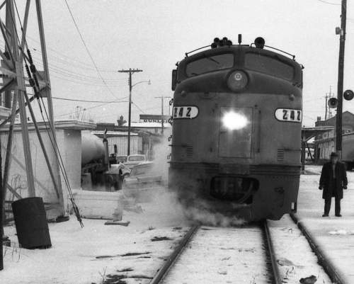 A vintage train approaches a snowy station, with steam rising and a person standing nearby.