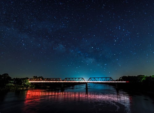 A starry night sky above a bridge reflecting in a calm river, with vibrant colors and a serene atmosphere.