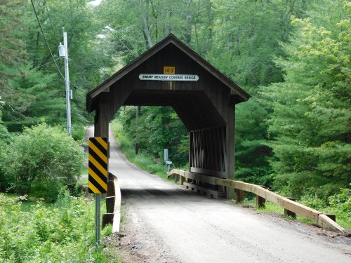 A wooden covered bridge spans a dirt road, surrounded by lush green trees and foliage.