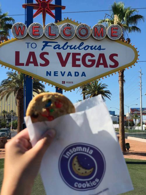 A hand holds a cookie in front of the iconic "Welcome to Fabulous Las Vegas" sign, with palm trees in the background.