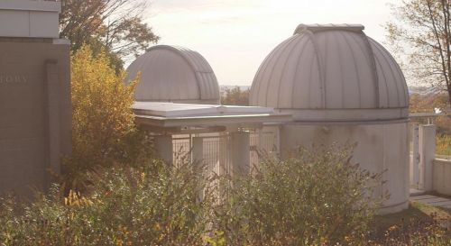 Two observatory domes surrounded by trees and foliage, with a soft morning light illuminating the scene.