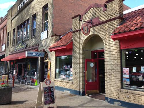 A brick storefront with a red awning, featuring a sign for a shop and nearby cafes in a quaint downtown area.