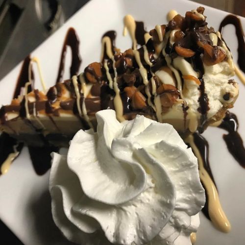 A slice of dessert topped with nuts and drizzled with chocolate and caramel, accompanied by a dollop of whipped cream.