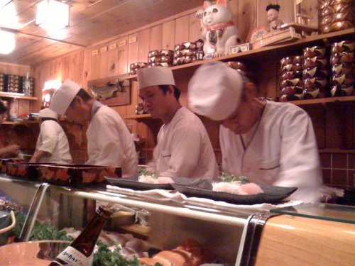 Three sushi chefs prepare dishes in a traditional Japanese restaurant, with wooden decor and various sushi on display.