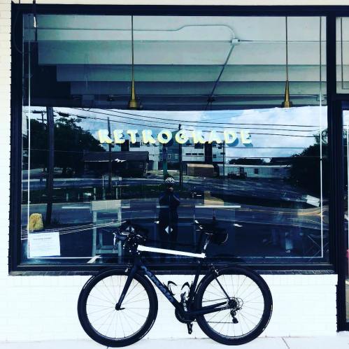 A black bicycle parked in front of a shop window displaying the sign "RETROGMADE" and a reflection of the street.