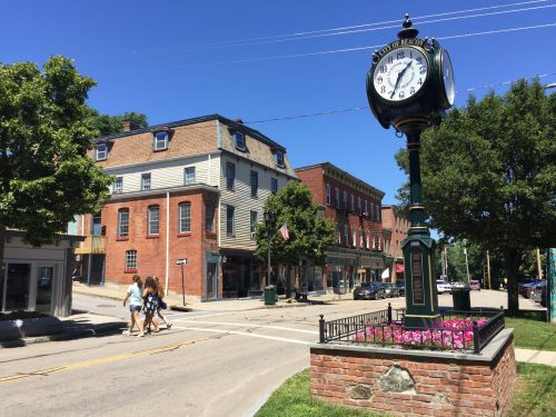 A historic town square featuring a clock tower, brick buildings, and pedestrians walking on a sunny day.