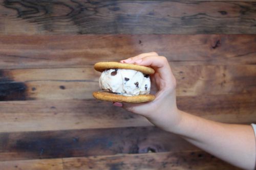 A hand holding a cookie sandwich filled with chocolate chip ice cream against a wooden background.