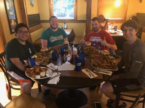 Four friends sit at a table filled with burgers, fries, and drinks, smiling and enjoying their meal together.