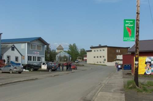 A small town street with buildings, parked cars, and people gathered, featuring a welcome banner for Dillingham.