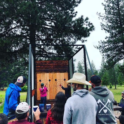 A group of people watches as a person throws an axe at a wooden target in a forested outdoor setting.