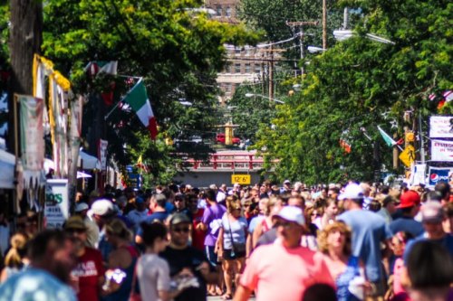 A bustling street fair filled with crowds, colorful banners, and food stalls under a sunny sky.