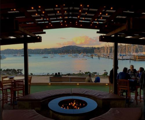 Cozy waterfront view from a restaurant with a fire pit, boats in the harbor, and a sunset over the hills.