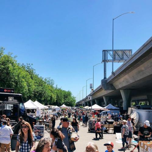 A bustling outdoor market scene with vendors, shoppers, and food trucks under a clear blue sky.