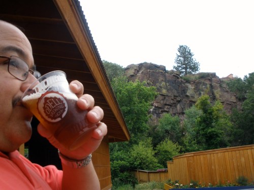 A person enjoys a beer while overlooking a rocky hillside and trees in a scenic outdoor setting.
