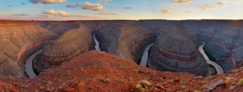 A panoramic view of a canyon with layered rock formations and a winding river at sunset, surrounded by clouds.