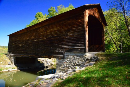 A rustic wooden covered bridge spans a calm stream, surrounded by green trees and a clear blue sky.