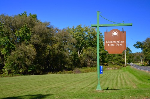 Sign for Glimmerglass State Park beside a grassy area and trees, with a clear blue sky in the background.