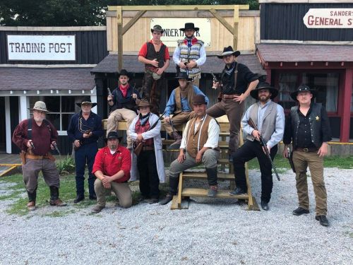 A group of men dressed in Western attire poses together in front of a rustic trading post and general store.