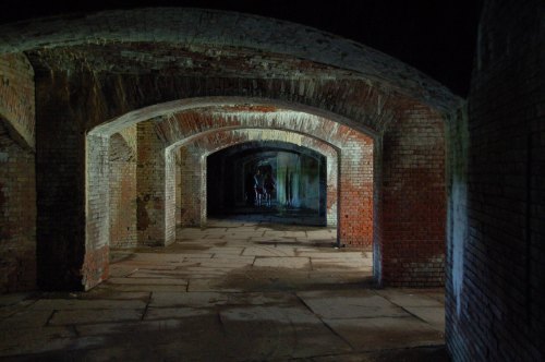 A dimly lit brick corridor with arched ceilings, leading to a shadowy figure in the distance.