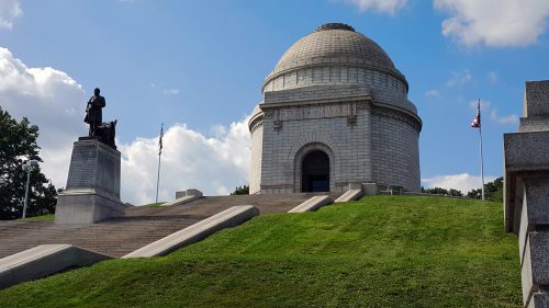 A large stone memorial with a dome, flanked by flags and a statue, set on a grassy hill under a blue sky.