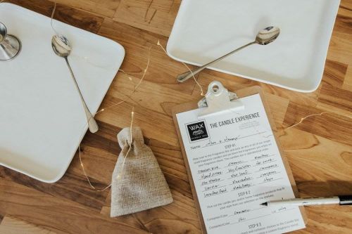 A wooden table with two white plates, spoons, a burlap bag, and a clipboard with a candle-making experience form.