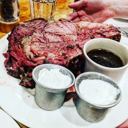 A large, juicy prime rib served on a plate with two cups of creamy sauce and a small bowl of gravy.