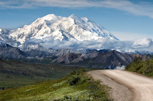 A winding dirt road leads through green hills, with a majestic snow-capped mountain in the background under a blue sky.