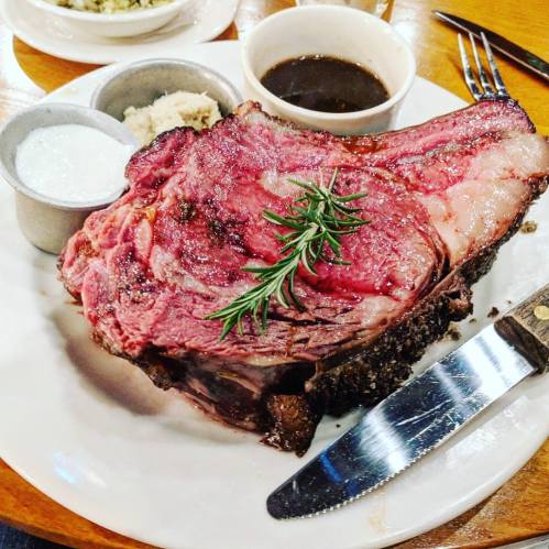 A large, juicy prime rib served with dipping sauces and a sprig of rosemary on a white plate.