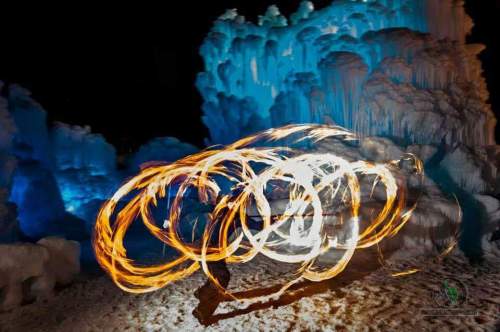 A performer spins fire in front of icy formations, creating dynamic light trails against a dark background.