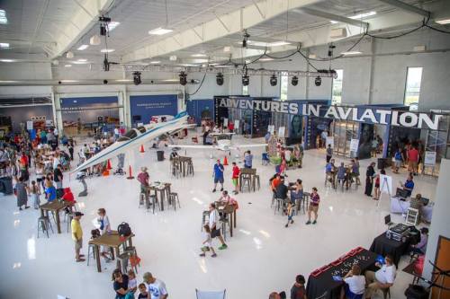 A spacious aviation event with people mingling, tables set up, and an airplane model hanging from the ceiling.