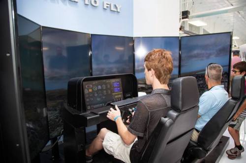 A person sits at a flight simulator with multiple screens, while another person observes nearby.