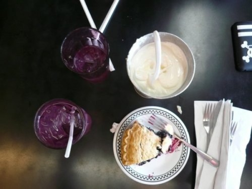 A table with two purple drinks, a bowl of ice cream, and a slice of blueberry pie on a black surface.