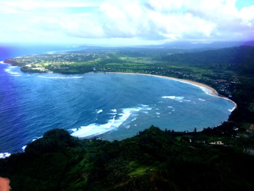 Aerial view of a coastal landscape featuring a curved beach, rolling waves, and lush green hills under a cloudy sky.