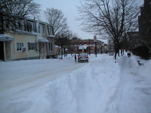 A snowy street scene with a car driving, lined by houses and trees, and pedestrians walking on the sidewalk.