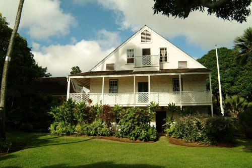 A two-story white house with a porch, surrounded by lush greenery and tropical plants under a partly cloudy sky.