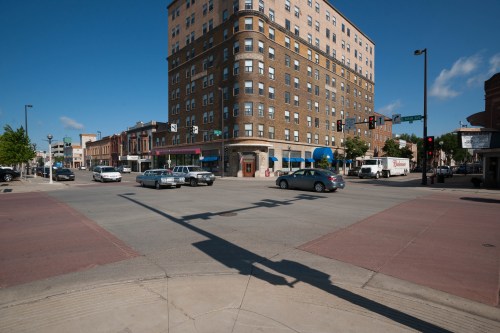 A busy city intersection with cars, a tall brick building, and clear blue skies. Traffic lights and shadows are visible.