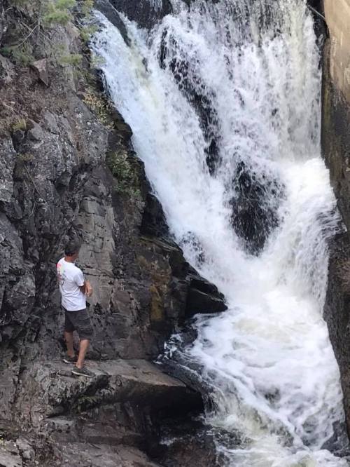 A person stands on rocky terrain beside a cascading waterfall, surrounded by nature.