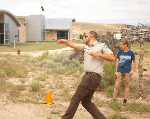 A park ranger demonstrates throwing a rope while a young boy watches in a desert landscape.