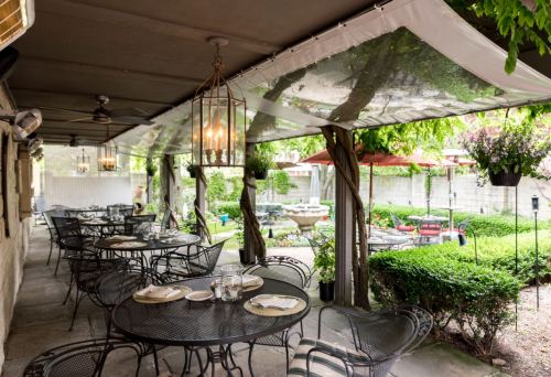 Outdoor dining area with metal tables and chairs, surrounded by greenery and a garden with a fountain in the background.